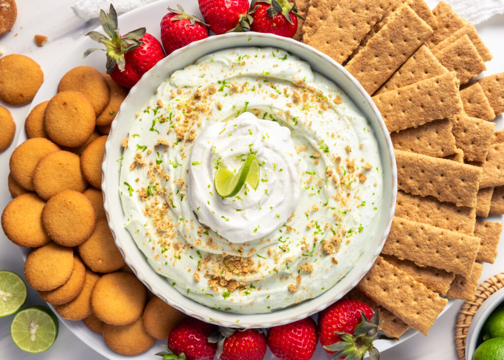 Key Lime Pie Protein Cheesecake Dip topped with whipped cream and served with graham crackers, Nilla wafers, and strawberries