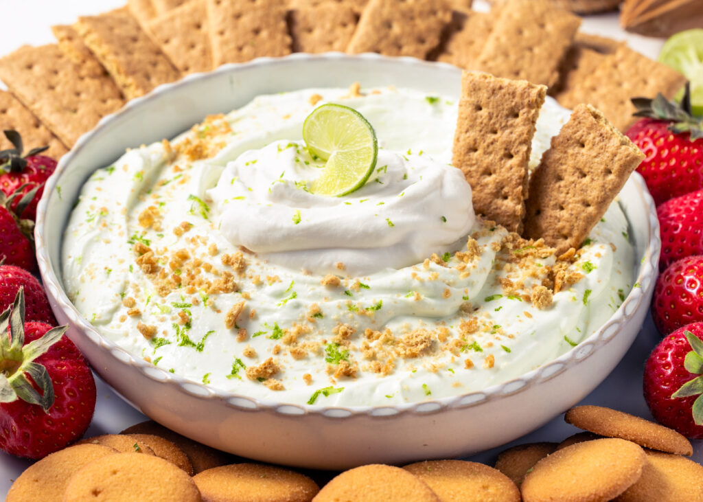 Key Lime Pie Protein Cheesecake Dip topped with whipped cream and served with graham crackers, Nilla wafers, and strawberries